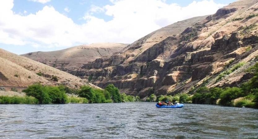 a raft is paddled by outward bound students on a calm river. There are high canyon walls surround them. 